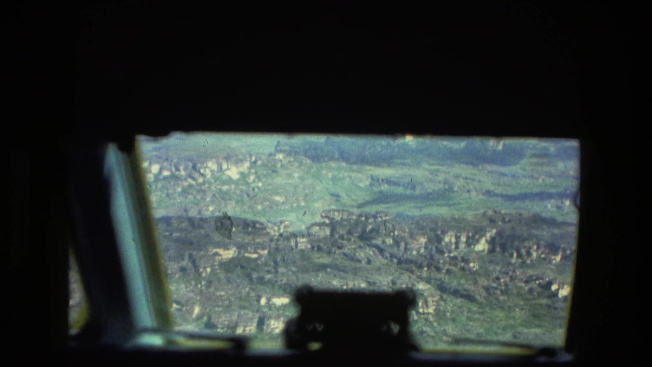 Helicopter window view over distant cliffs and forest, framed by dark cockpit silhouettes.