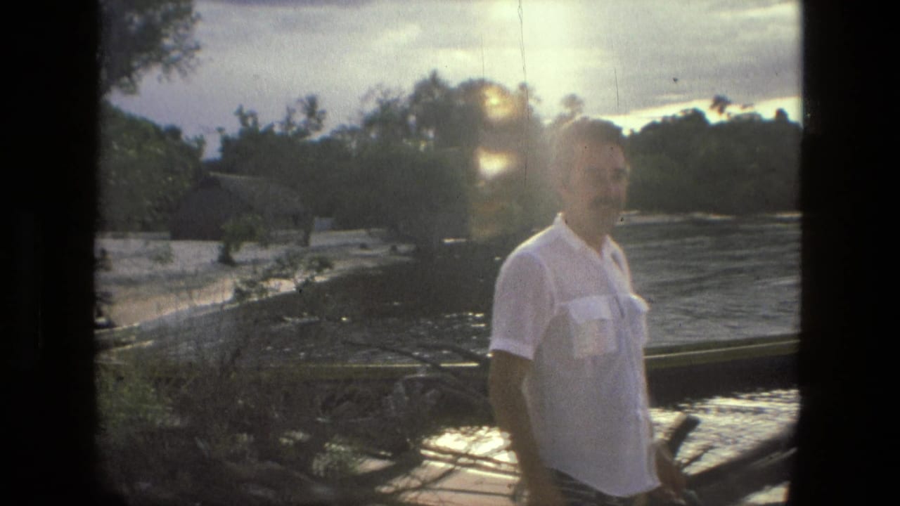 Man in a white shirt stands near a riverbank with boats and trees behind him.