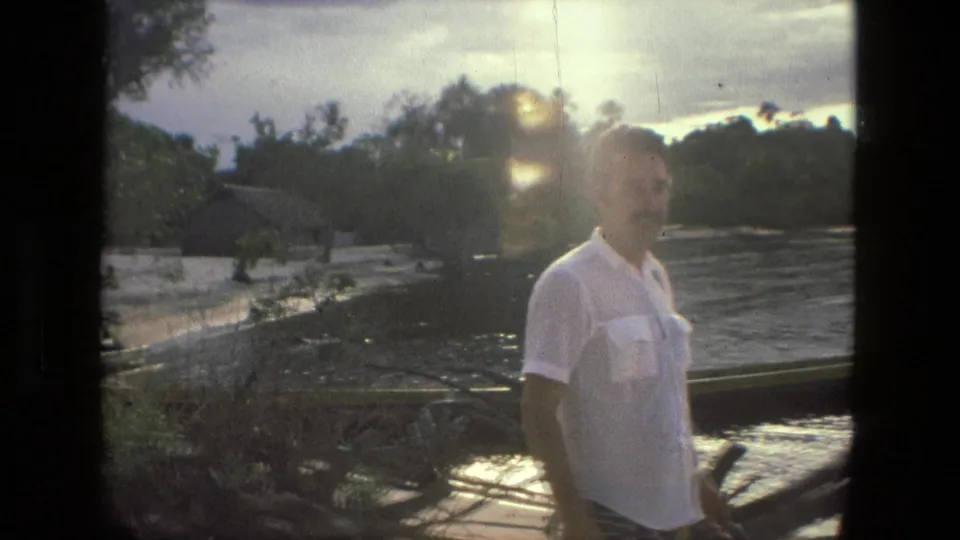 In 1979 Manaus home-movie footage, a man in a white shirt stands beside a sandy riverside with wooden boats, trees, and water behind him.