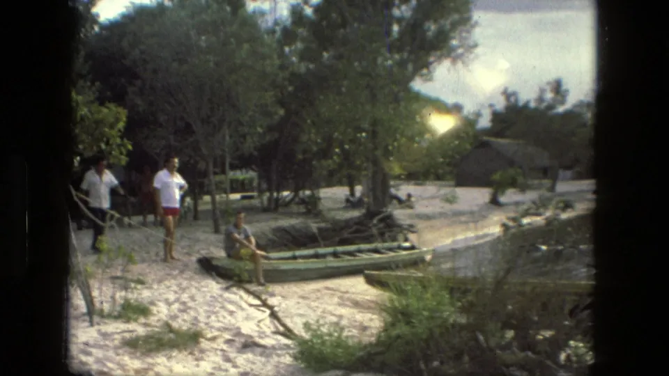 Adults appear near wooden boats on a sandy riverside in Manaus, with trees, water, and a thatched structure visible in the background.