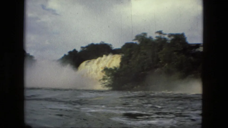 A broad waterfall drops beside dense jungle into turbulent water, with mist rising across the river surface.