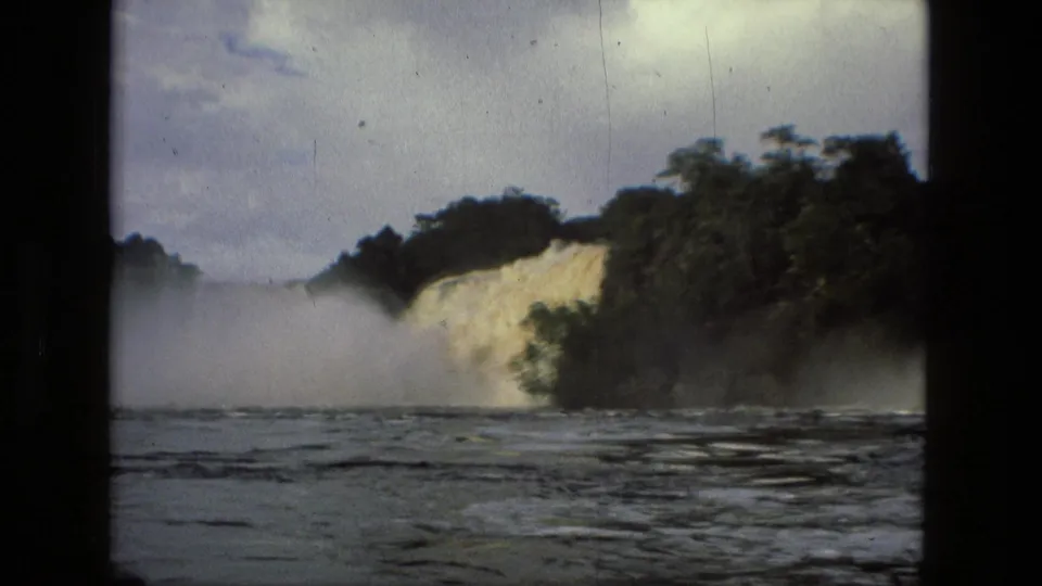 The waterfall continues across the frame, with jungle above the drop and rippled river water in the foreground.