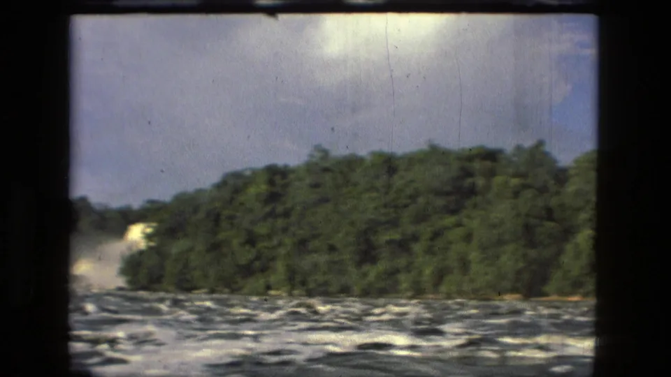 1979 Manaus, Brazil 8mm frame showing choppy water, dense green trees, pale distant spray, and film scratches.