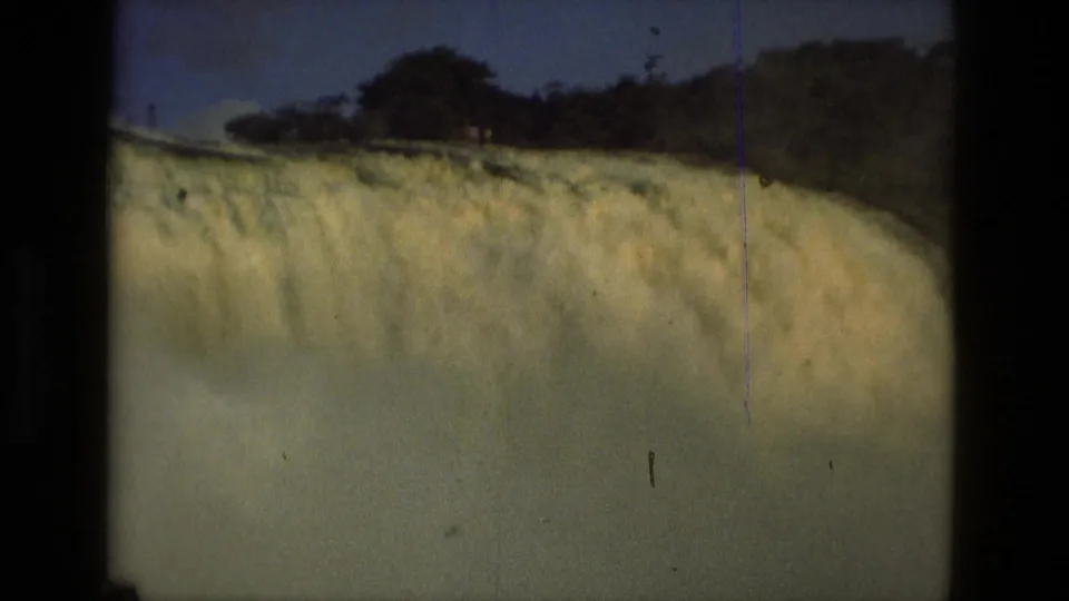 1979 8mm view near Manaus, Brazil, showing the waterfall crest, dense mist, treeline, and small distant figures near the top edge.