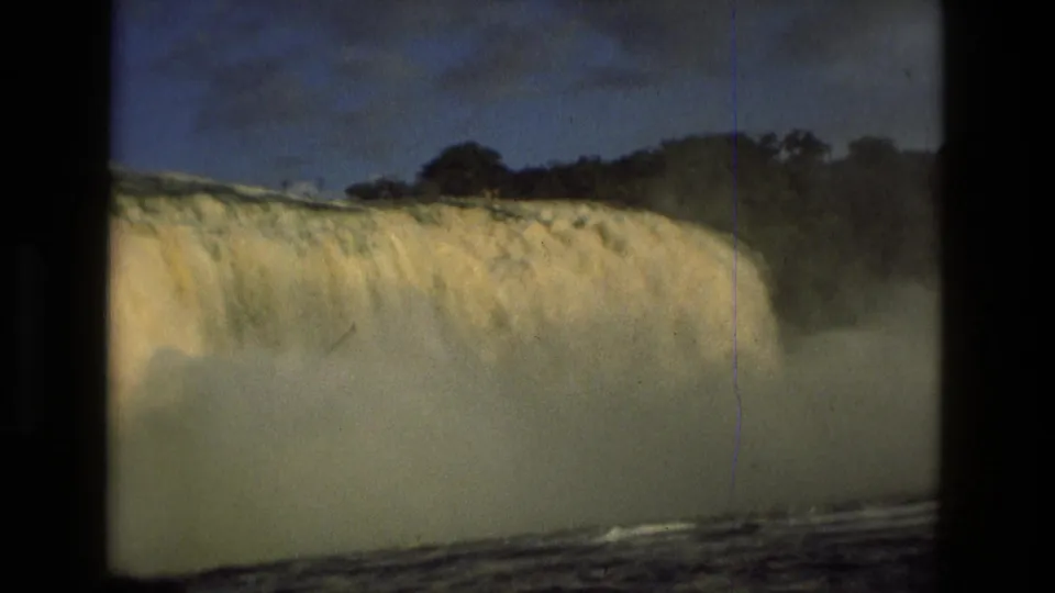 1979 8mm frame near Manaus, Brazil, showing a broad curtain of falling water, mist, treeline, and river surface below.