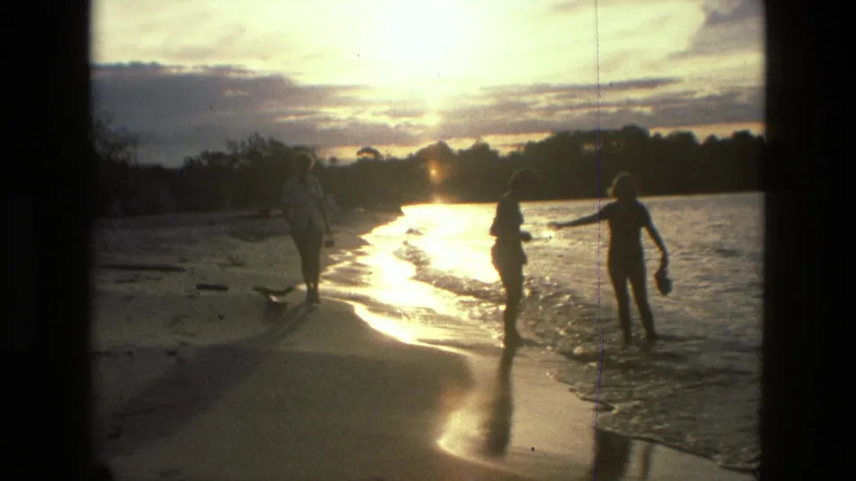 1979 Manaus home-movie frame of three distant people silhouetted along a sandy shoreline beside sunlit water.