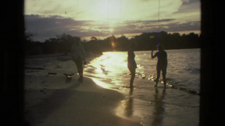 A 1979 8mm beach scene in Manaus with distant figures, wet sand, small waves, and strong sunset glare.
