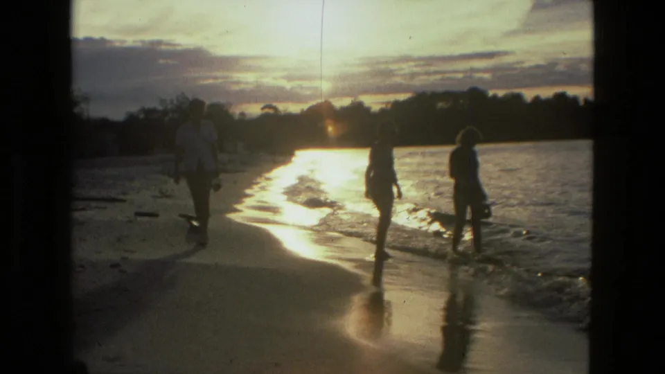 1979 Manaus 8mm frame of silhouetted people walking at the edge of shallow water, with sun glare and trees beyond.