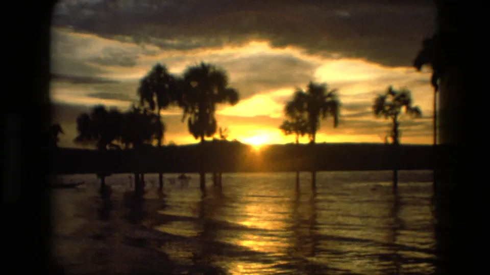 1979 8mm sunset frame showing water reflections and dark palm shapes along a Manaus shoreline.