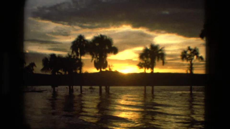 8mm home-movie frame from Manaus, Brazil in 1979, with a low sun, silhouetted palms, and shimmering water in warm dusk light.