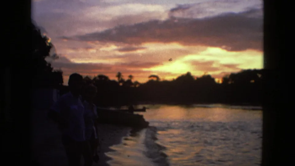 1979 Manaus footage frame with silhouetted people beside the river, a dark treeline, and bright sunset clouds reflected on the water.