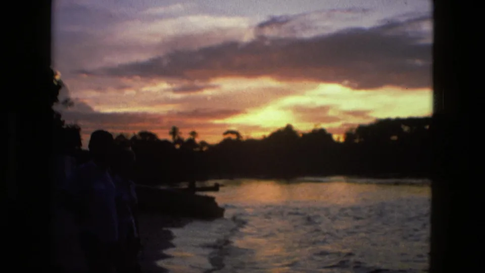 1979 Manaus 8mm frame showing a tropical river sunset, dark shoreline figures, water reflections, and tree silhouettes across the horizon.