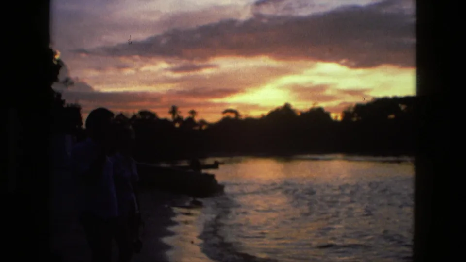 1979 Manaus home-movie frame with a couple in silhouette by the river, sunset clouds overhead, and reflected light on the water surface.