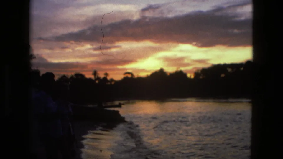 1979 Manaus home-movie frame with a silhouetted person at the riverbank as sunset color spreads over the water and treeline.
