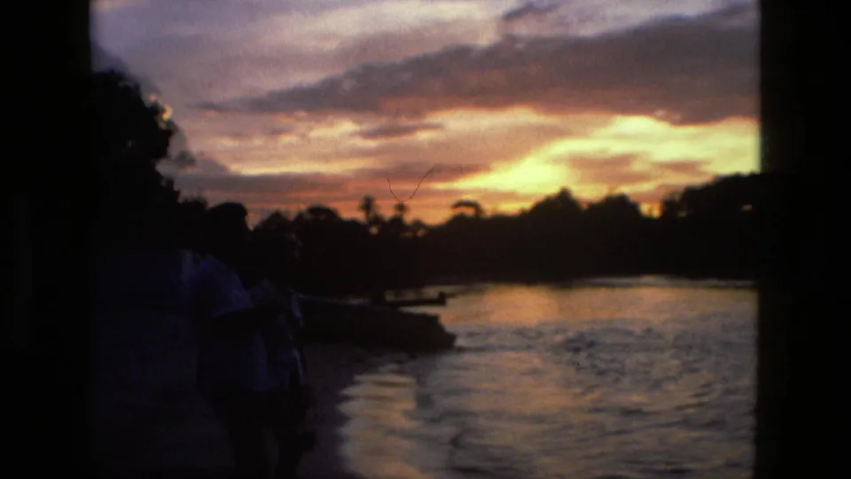 Manaus, Brazil 8mm home-movie frame showing two shadowed friends near the water as the sunset glows behind palm silhouettes.
