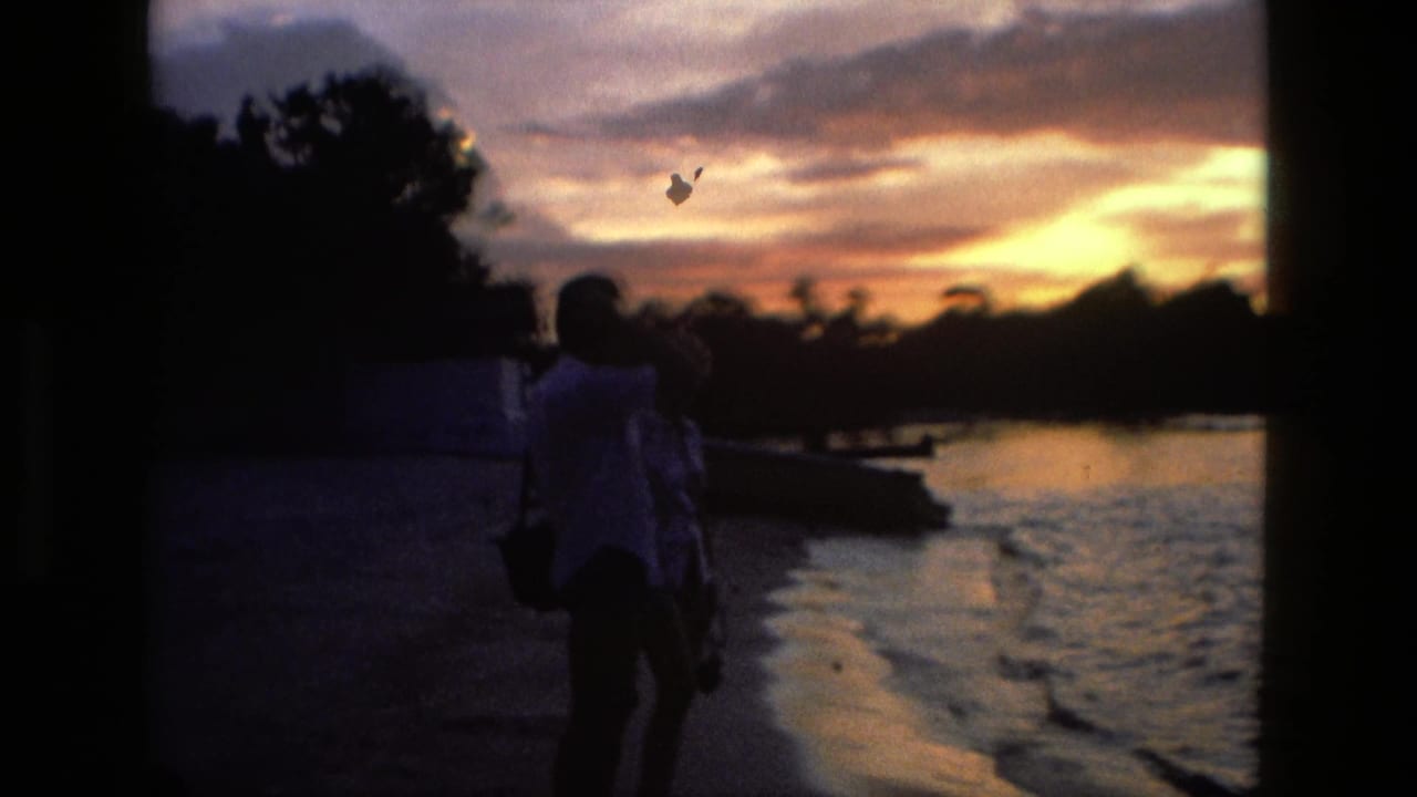 Silhouetted couple stands near a river at sunset with trees and glowing clouds behind them.