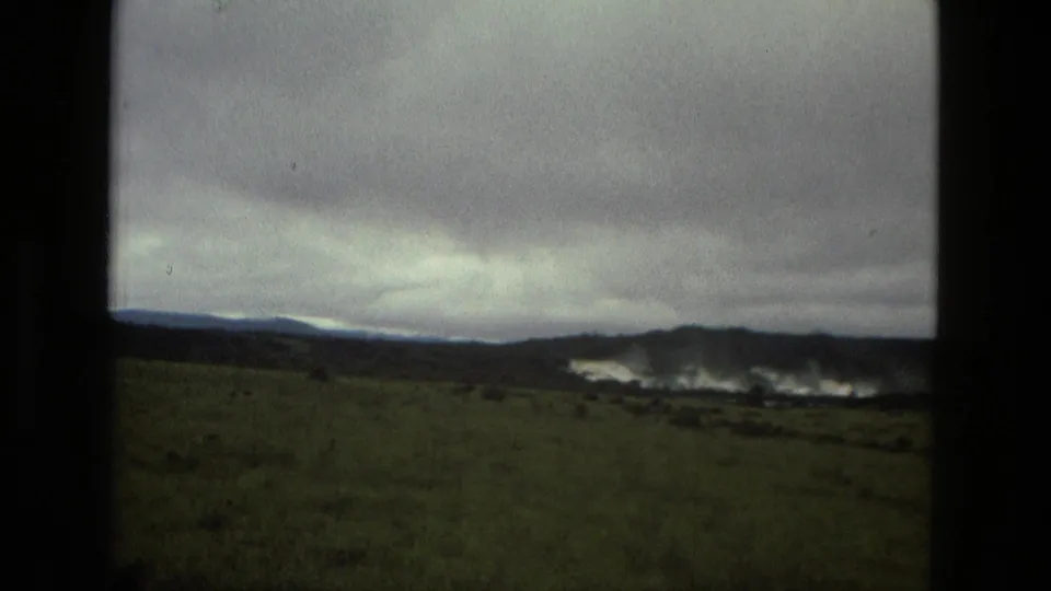 1979 8mm Manaus landscape frame with a grassy foreground, low hills, cloud cover, and light haze or water in the distance.