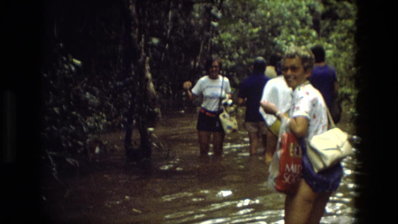 A woman smiles toward the camera while tourists wade through shallow water behind her.