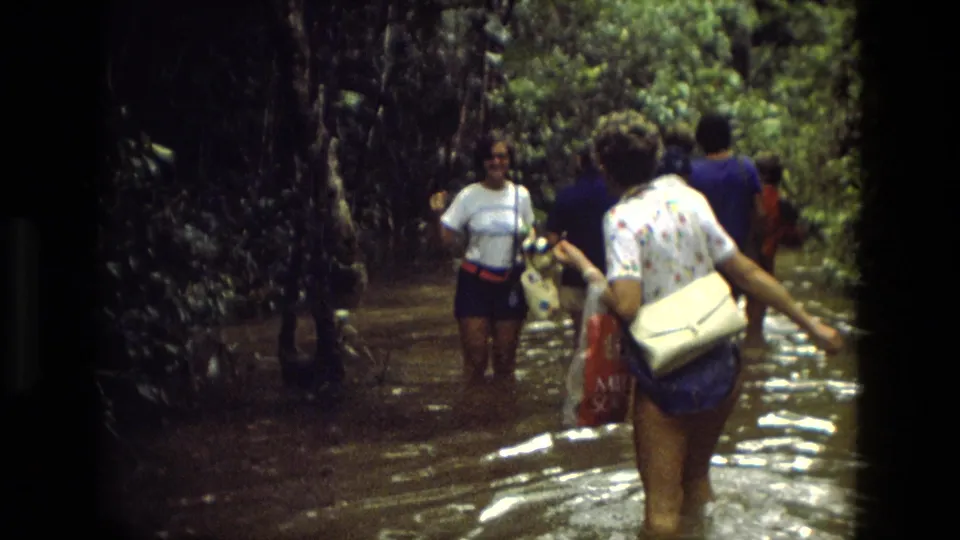 1979 Manaus home-movie frame showing adults walking through brown floodwater beneath heavy tropical vegetation.