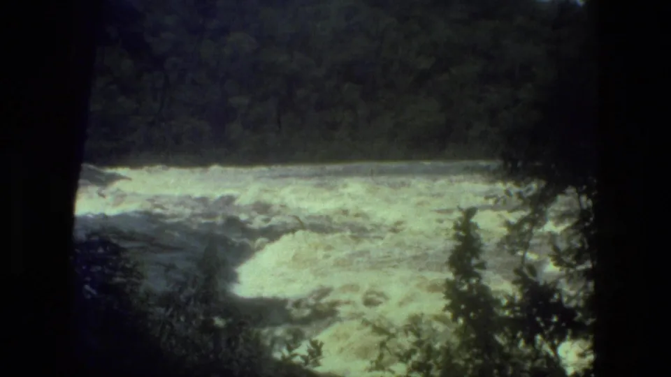 1979 8mm Manaus, Brazil footage showing turbulent river rapids framed by dark tree trunks and rainforest leaves.
