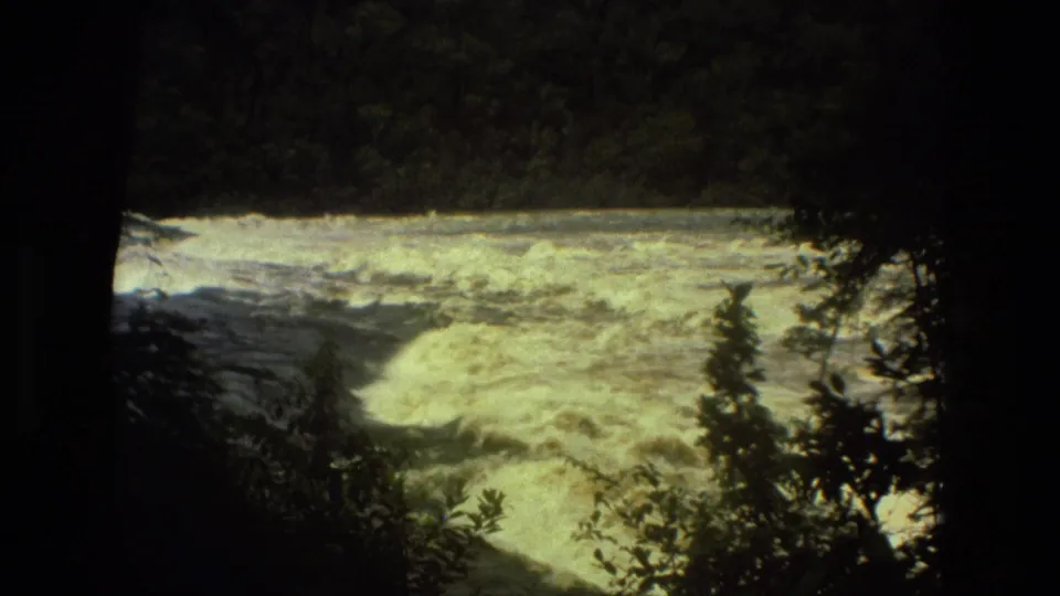 1979 8mm Manaus, Brazil frame with bright foaming rapids beyond shadowed rainforest branches and trees.