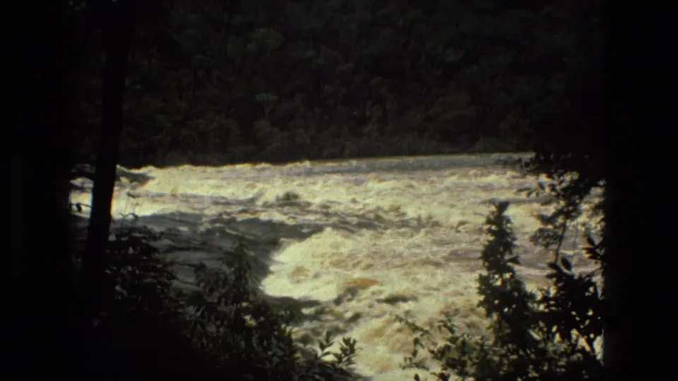1979 8mm Manaus, Brazil view of pale turbulent river water partially obscured by shaded rainforest vegetation.