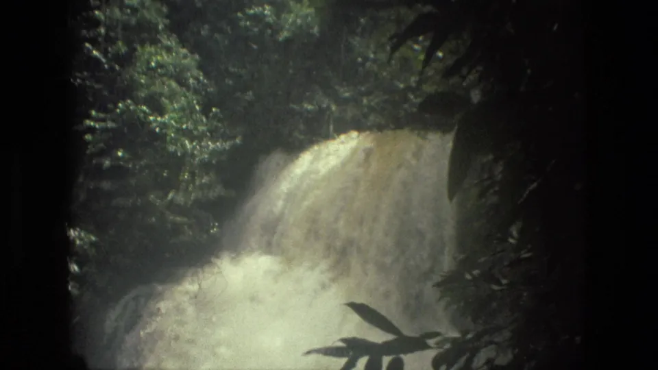 Closer 1979 8mm view near Manaus, Brazil, showing a broad waterfall, foaming water, and foreground jungle leaves.