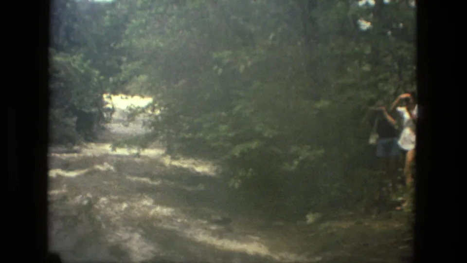 1979 8mm Manaus riverbank view with two people beside rushing water, one appearing to photograph the scene.