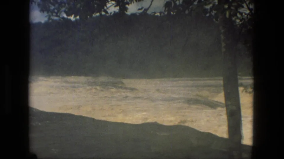 1979 8mm frame from Manaus, Brazil, showing foamy river rapids, a dark riverbank edge, forested hills, and a tree trunk in the foreground.