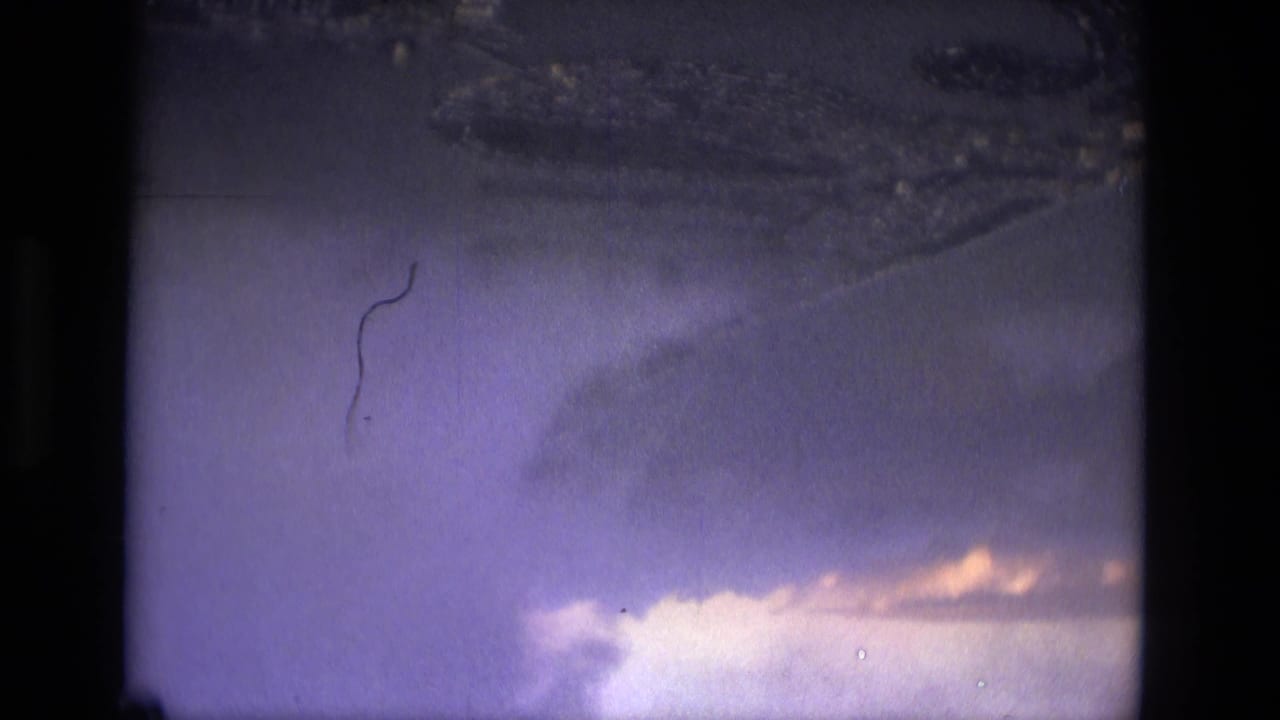 Clouded aerial view with dark land or water forms below and visible 8mm frame edges.