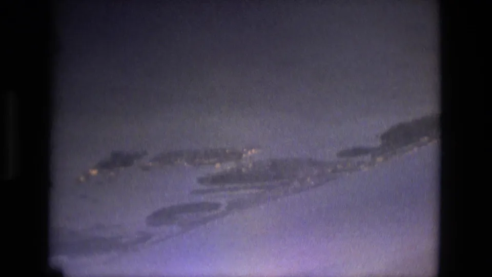 1979 8mm aerial frame, possibly near Manaus, with blue water, cloud cover, and scattered lights along an island or shoreline.