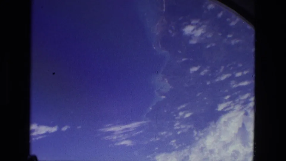 1979 8mm frame with blue atmosphere, scattered white cloud streaks, haze, and rounded window or gate edges around the view.