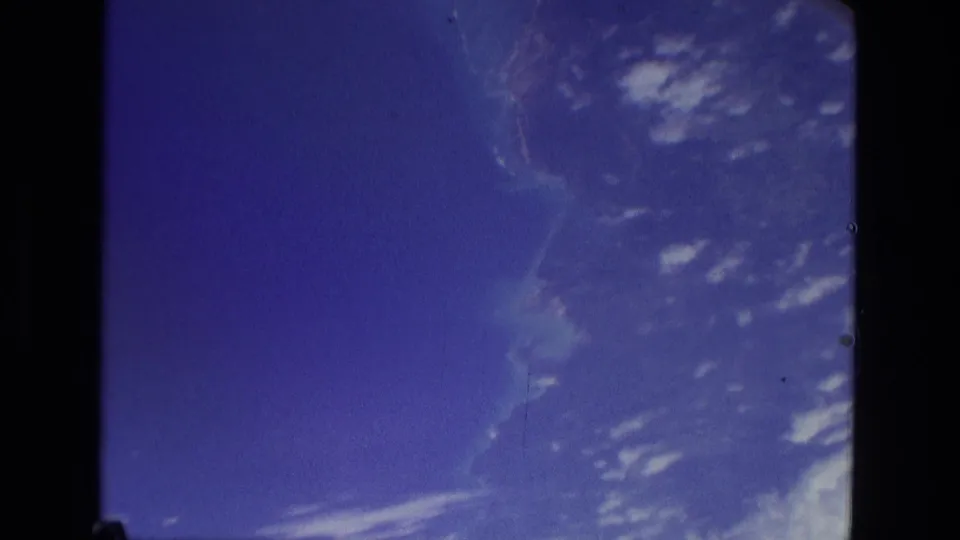 Late-1970s 8mm frame showing blue haze, scattered clouds, and a faint curved land or atmospheric shape near the window edge.
