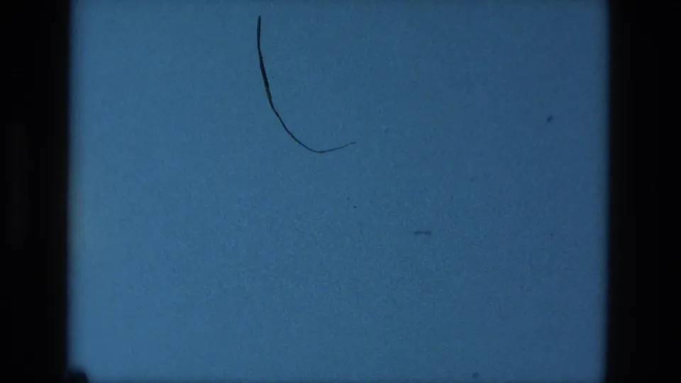 1979 Manaus 8mm frame of a blue screen-like surface with dark film borders, specks, and a curved hair or scratch mark near center right.