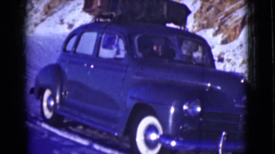 The 1940s-style blue sedan appears closer in frame on the snowy Alaska road, showing whitewall tires, roof luggage, and people inside.