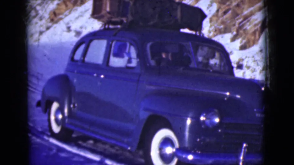 A close frame shows the blue 1940s sedan on a snowy Alaska mountain road, its rounded body, front headlight, whitewall tire, and roof load visible.