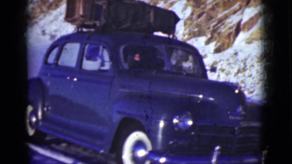 The blue vintage sedan continues past the camera on the snowy Alaska road, with roof luggage, whitewall tires, and occupants visible through the side windows.