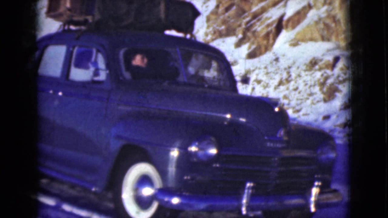 Blue vintage sedan with roof luggage on a snowy road beside rocky banks.