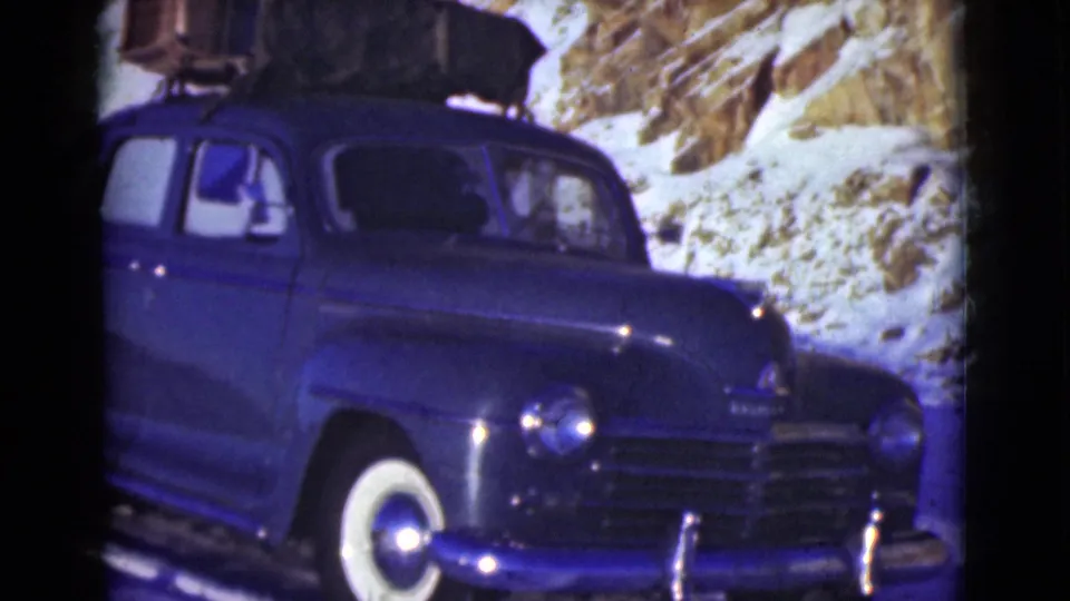 Blue mid-century sedan with luggage on top appears on a snowy Alaska road, with a person visible inside.