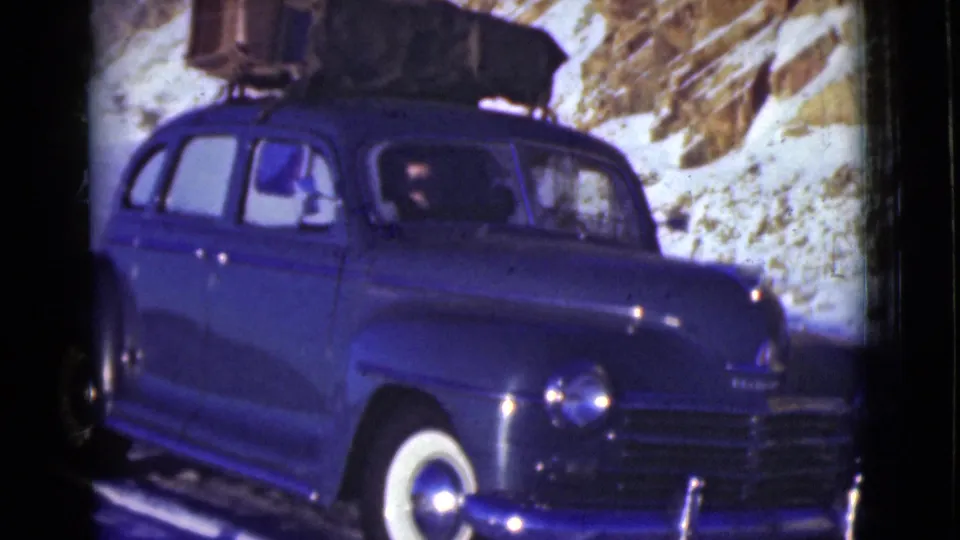 A blue 1940s sedan fills the frame as it travels along a snowy road in Alaska, with roof luggage visible.