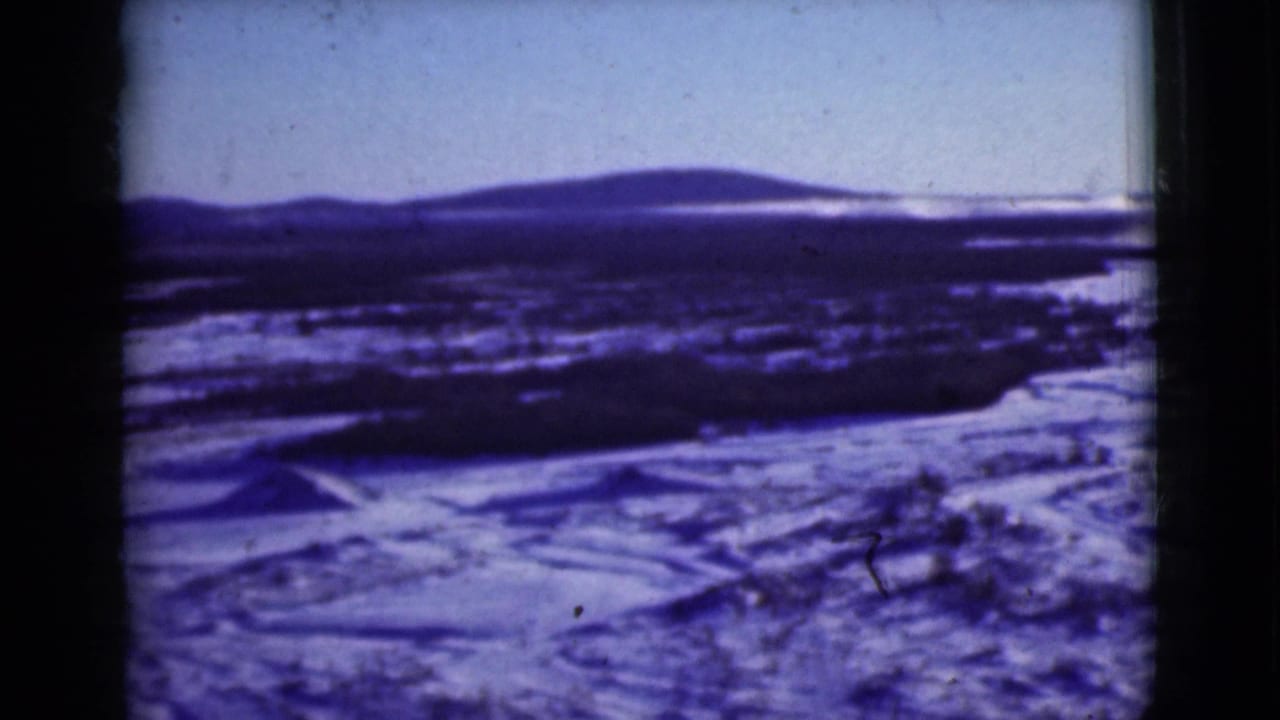 1946 8mm frame of an Alaskan snowfield, low dark ridges, and distant mountains with a blue-purple color cast.