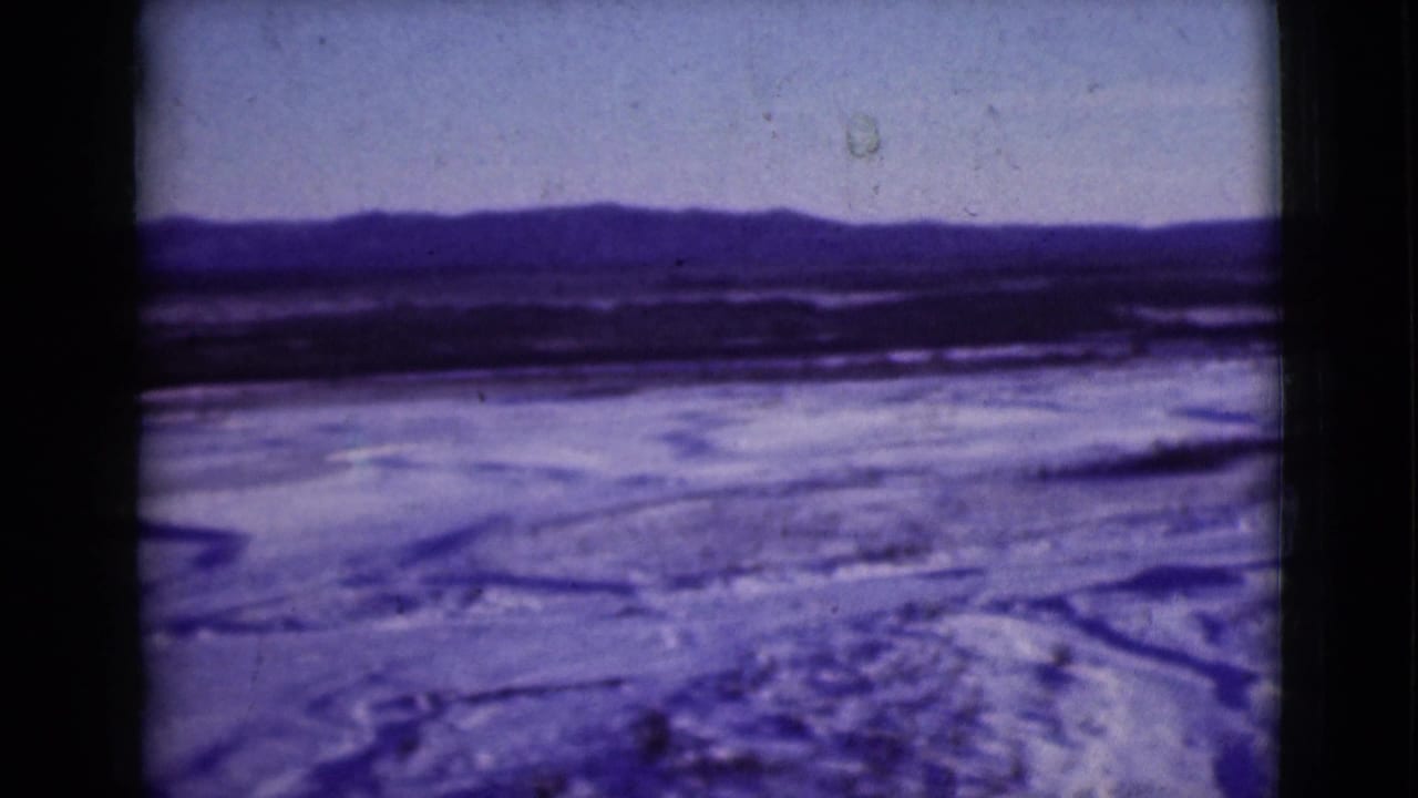 1946 Alaska home-movie frame of open winter ground, snow patches, possible ice channels, and a low mountain horizon.