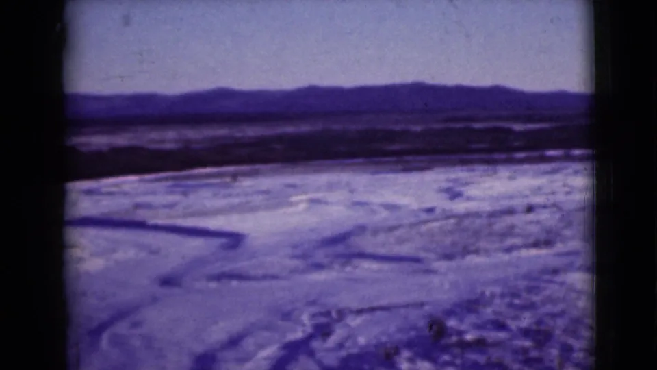 1946 Alaska 8mm still showing snowy open land, a dark channel across the foreground, and distant low mountains.