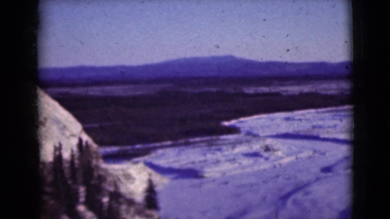 1946 Alaska home-movie frame with a snowy slope and dark pines overlooking a frozen river valley.