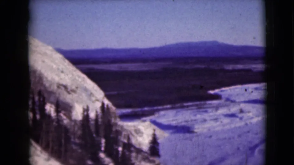 1946 Alaska 8mm frame showing snow-laden slope, pines, frozen valley floor, and distant mountains.