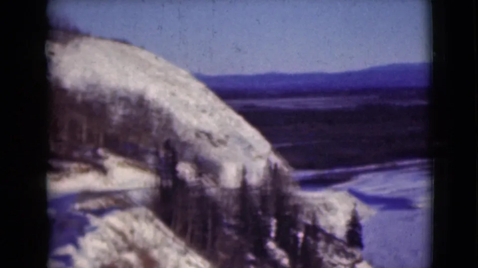 1946 Alaska home-movie view of a snowy bluff and pine trees above a frozen river plain and low ridges.