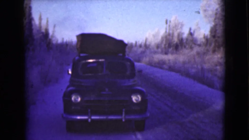 1946 Alaska home-movie frame of a dark vintage car carrying roof luggage on a narrow road bordered by winter trees.