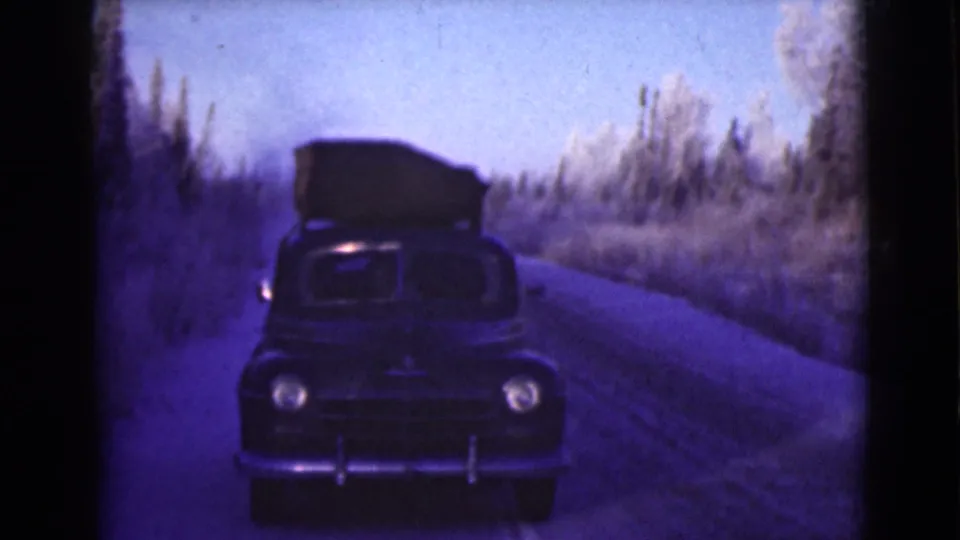 1946 Alaska frame showing a dark mid-century car with luggage on top moving toward the camera on a winter roadside.