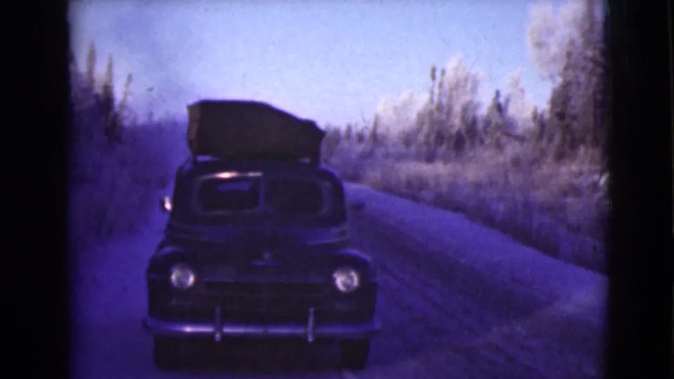 1946 Alaska 8mm frame with a roof-loaded vintage car centered on a narrow road through snow-dusted roadside trees.