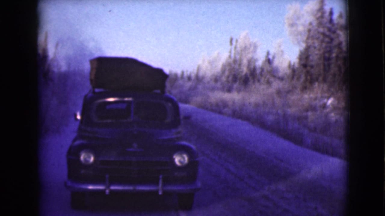 Dark vintage sedan approaches on a curving rural road beside snow-dusted trees.